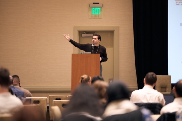 A speaker gesturing toward the screen during a talk